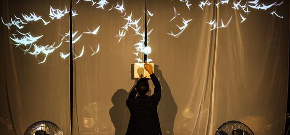 A photo of a stage, with black sheets draped as a backdrop to a projection of white birds and warm yellow light. On the stage is a man with his back t
