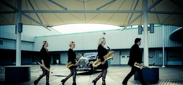 4 musicians carying various sizes of saxophone walking across a hangar building with a Rolls Royce in the background.