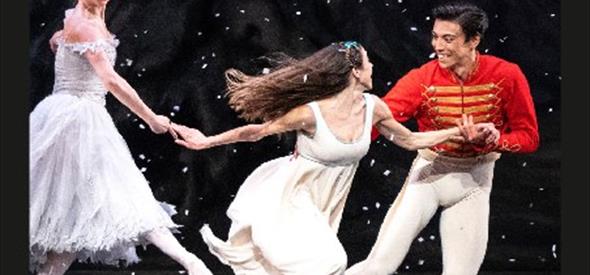 Three people on stage, two women wearing white dresses and the man holding one womens hand wearing a red top and white trousers