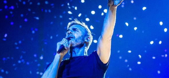 Gary barlow points to the sky while singing infront of a blue background