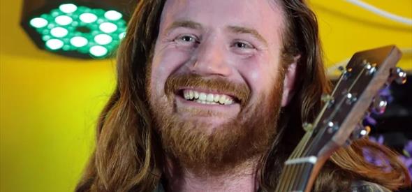 Close up of long haired male holding his guitar wearing a check shirt with a yellow background