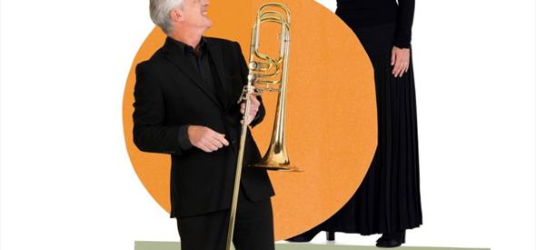 Image of a woman and a man both dressed in black against a white and orange background.  the man is holding a trombone.