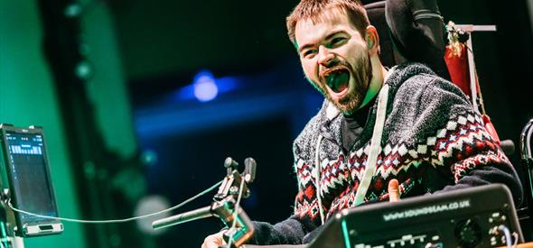 Man with beard sat in wheelchair laughing heartily with mouth open under theatre lights