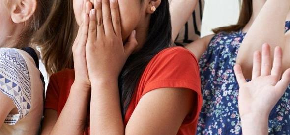 A child wearing a coral red top is acting surprised by covering their mouth with their hands with a wide-eyed expression. They are surrounded by other