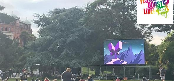 Group of people sitting on the grass in the gardens looking a large screen