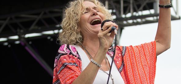 Female singer with blond curly hair singing into a microphone, wearing a red jacket and with her left hand up in the air