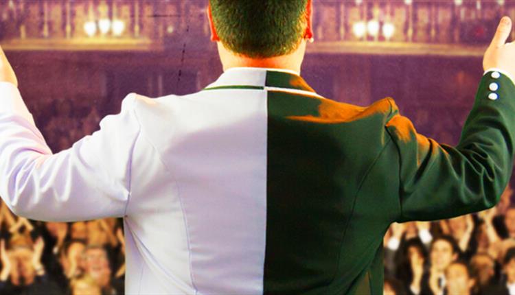 A man standing with his back to the camera with his hands up in the air, wearing a half black, half white blazer. An  audience of people behind him.