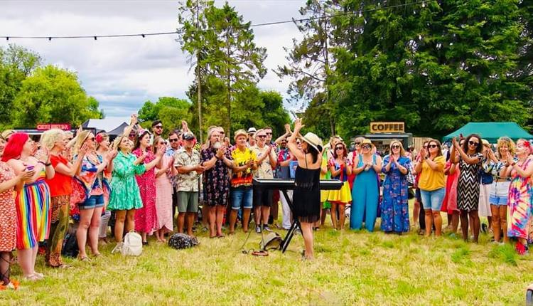 Semi circle of people dressed in brightly coloured clothes singing with a conductor in front of a keyboard