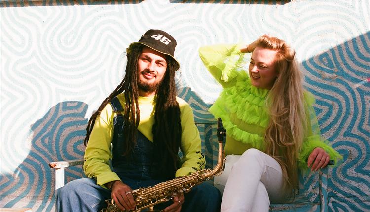 Blue and white swirly background with a man with dreadlocks and a woman in a brilliant green feathery top sitting. Man is holding a saxophone  and loo