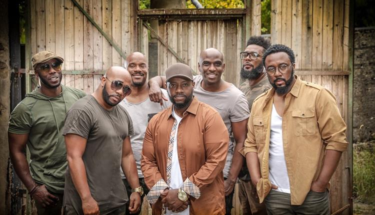 Seven men in earthy toned clothing and a wooden fence with trees in the background