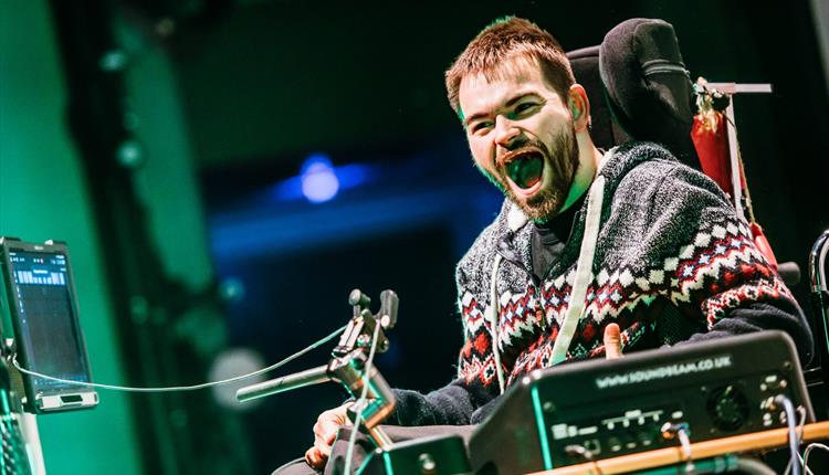 Man with beard sat in wheelchair laughing heartily with mouth open under theatre lights
