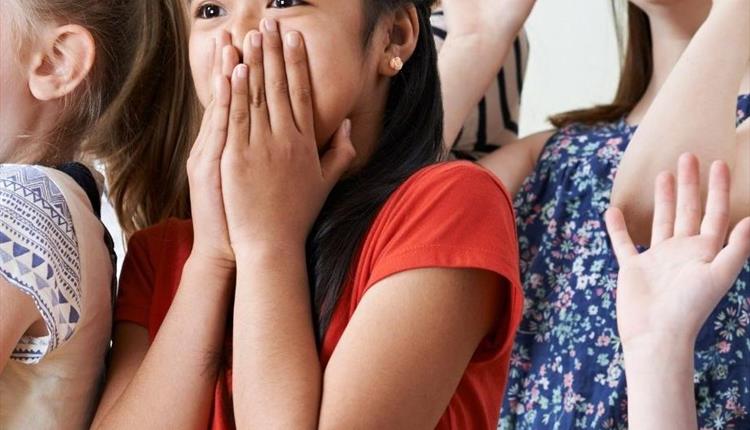 A child wearing a coral red top is acting surprised by covering their mouth with their hands with a wide-eyed expression. They are surrounded by other