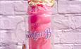 A vibrant pink jammy-dodger flavoured milkshake held infront of an equally bright pink brick wall.