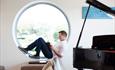 Man reading a book, sat in a circular window sill with grand piano in foreground.